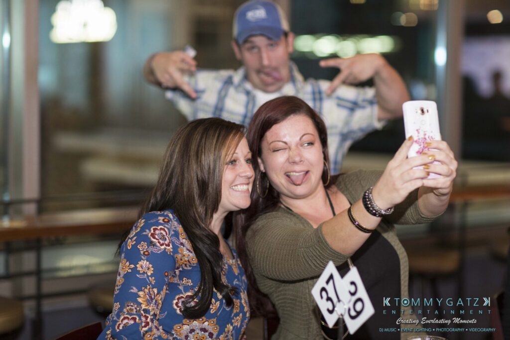 18A0139 - TOMMY GATZ ENTERTAINMENT Two women are taking a selfie at a table with a 37.9 sign, while a man in the background makes a playful face and gesture.