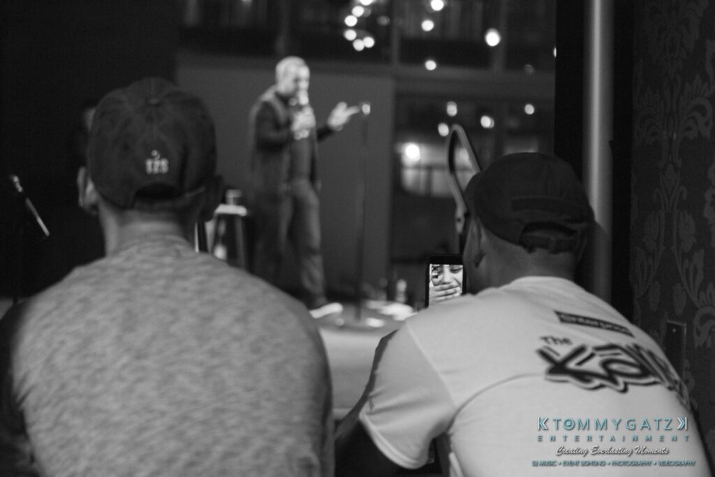 18A0146A-1 - TOMMY GATZ ENTERTAINMENT Two people seated in the foreground of an indoor event, one recording a blurred speaker on stage with a smartphone.