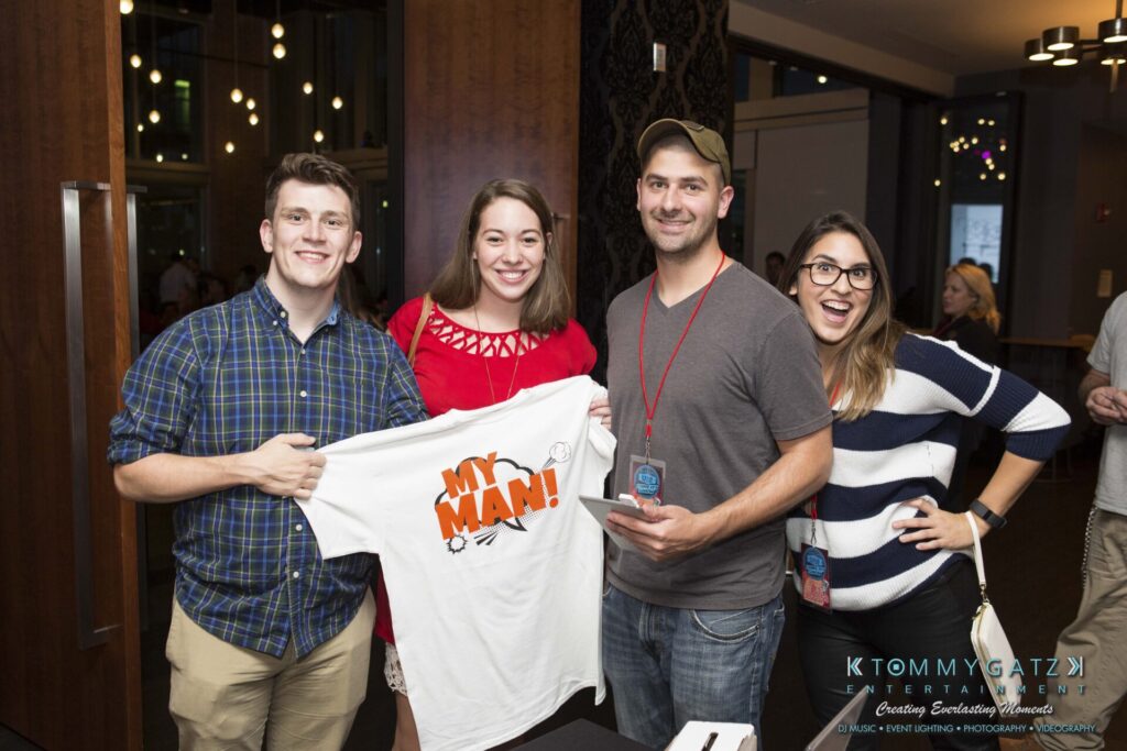 18A0153 - TOMMY GATZ ENTERTAINMENT Four people smiling at the camera; one holds a T-shirt with "MY MAN!" printed on it. They stand indoors at an event, wearing casual clothing and event lanyards.