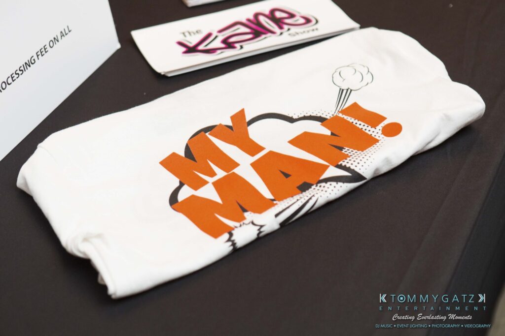 18A0164 - TOMMY GATZ ENTERTAINMENT A folded white shirt with "My Man!" printed in bold orange and black letters lies on a table near promotional cards.