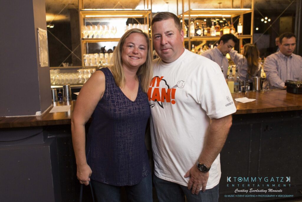 18A0250 - TOMMY GATZ ENTERTAINMENT A man and woman stand together in front of a bar, smiling at the camera. Other people are visible in the background.