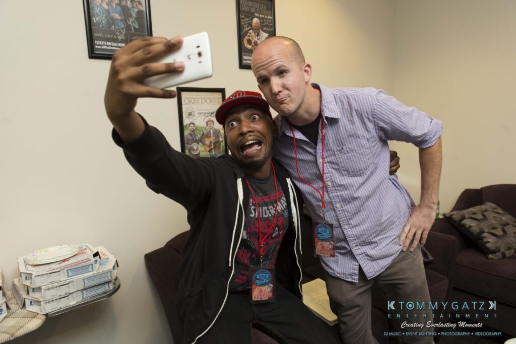 18A0290 - TOMMY GATZ ENTERTAINMENT Two men taking a selfie in a room with pizza boxes on the side table. One is wearing a black jacket and red cap, and the other is wearing a striped shirt. Posters are on the wall behind them.