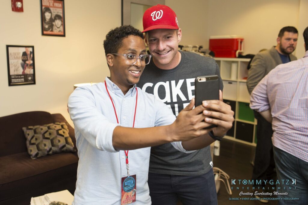 18A0293 - TOMMY GATZ ENTERTAINMENT Two smiling men taking a selfie together in a room with casual decor. One is wearing a red cap and gray shirt, the other has glasses and a lanyard.