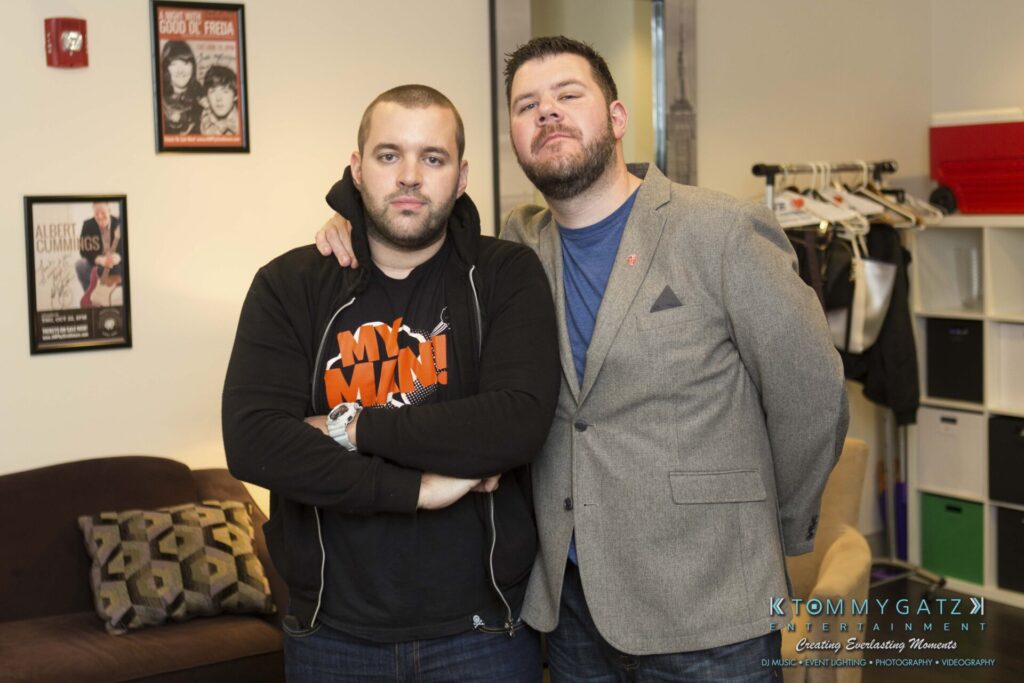 18A0303 - TOMMY GATZ ENTERTAINMENT Two men stand together in a room, one with arms crossed. The room has posters, a couch with cushions, and a clothing rack.
