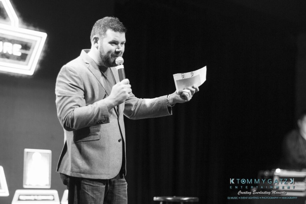 18A0331 - TOMMY GATZ ENTERTAINMENT Man holding a microphone and papers, speaking on stage in a black and white setting.