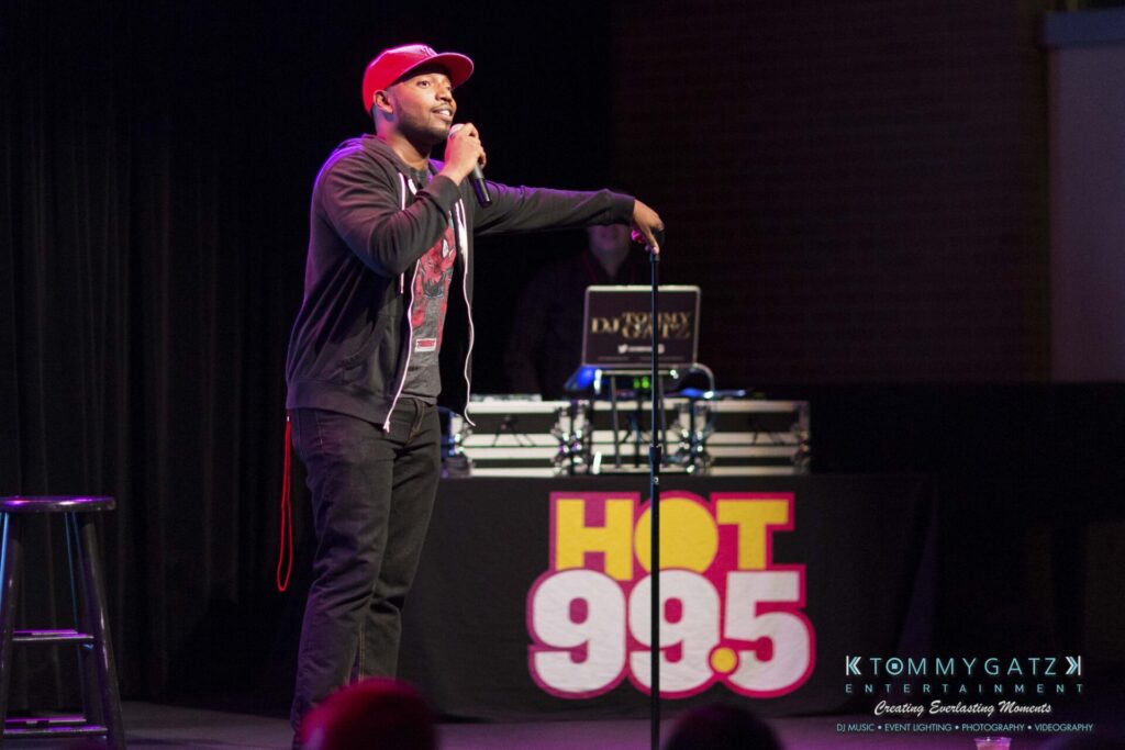 18A0411 - TOMMY GATZ ENTERTAINMENT A person on stage performing stand-up comedy, holding a microphone. A DJ booth with "HOT 99.5" is visible in the background.