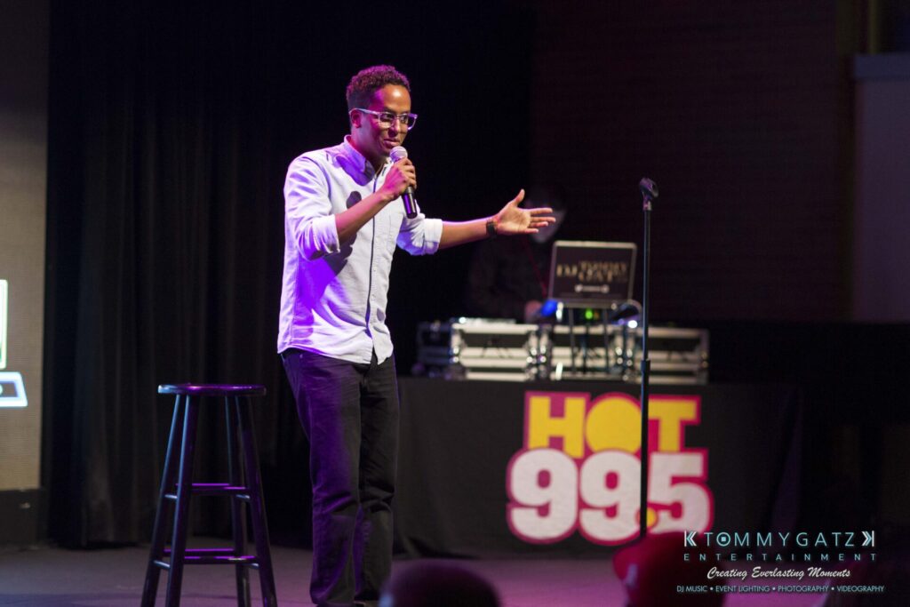 18A0460 - TOMMY GATZ ENTERTAINMENT A person performs stand-up comedy on stage with a microphone and a stool. A DJ booth with "Hot 99.5" is visible in the background.
