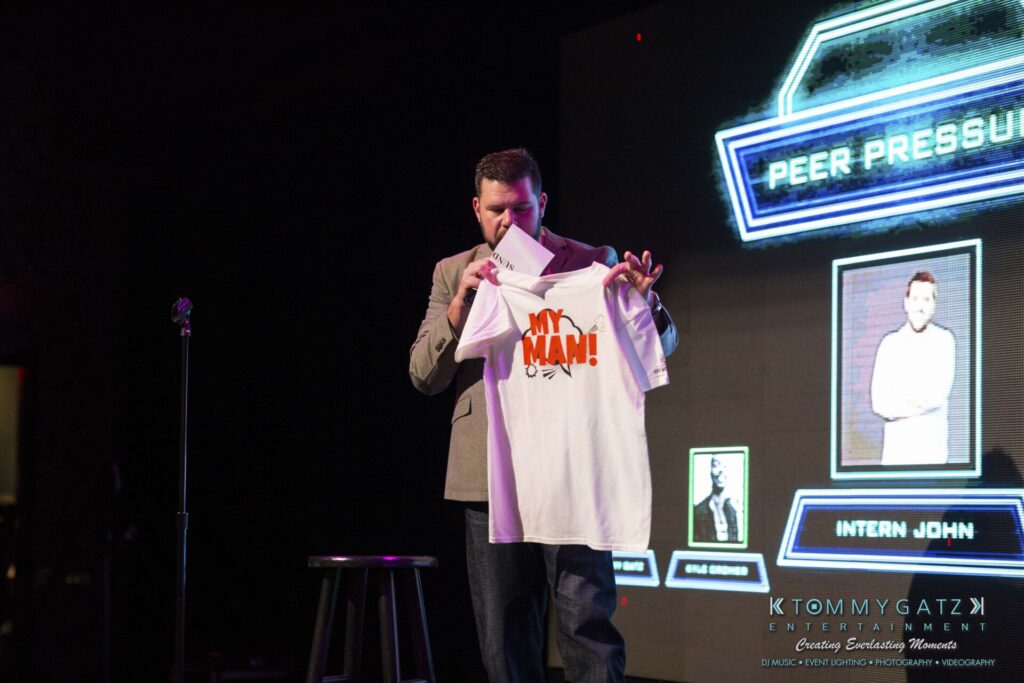 18A0567 - TOMMY GATZ ENTERTAINMENT A man stands on stage holding a white T-shirt with the words "MY MAN!" printed on it. A microphone stand and a screen displaying "PEER PRESSURE" are in the background.