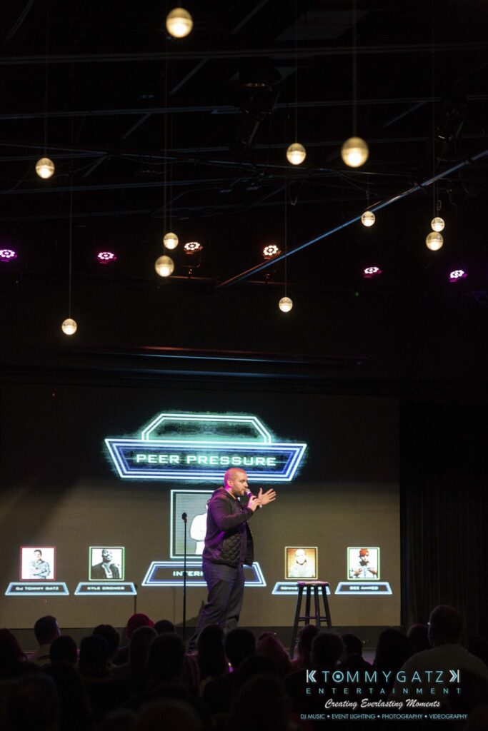 18A0669 - TOMMY GATZ ENTERTAINMENT A person on stage presents a game show titled "Peer Pressure" with a futuristic backdrop and illuminated hanging lights above.