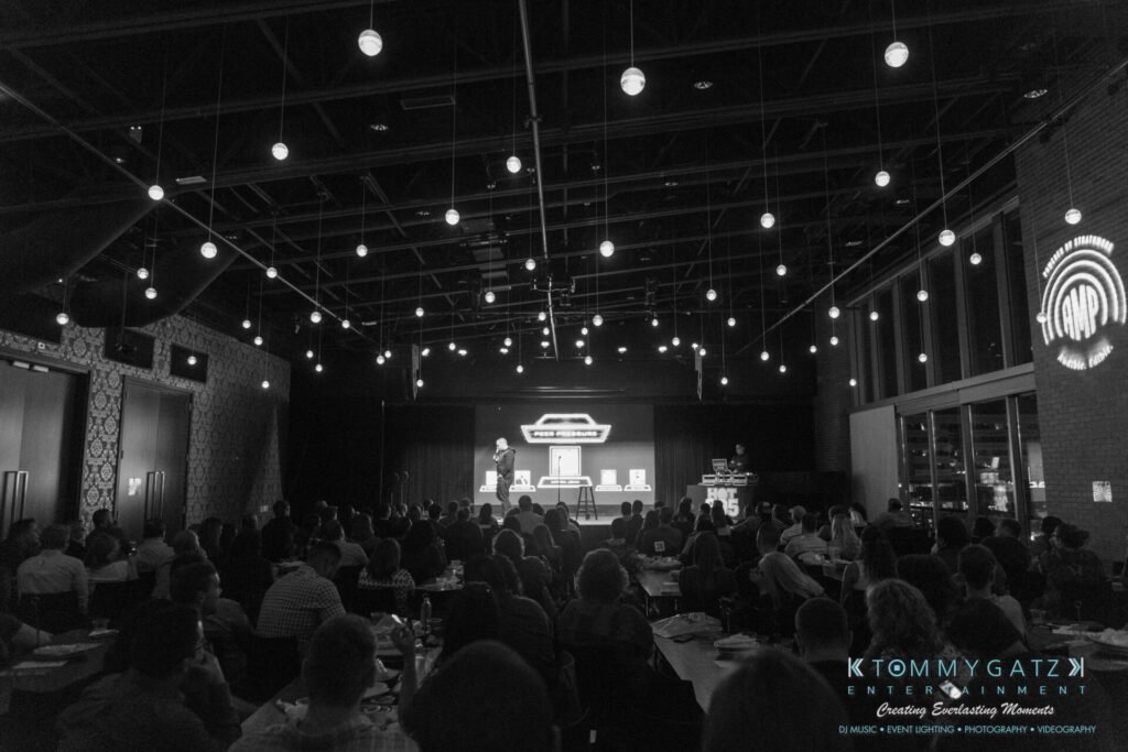 18A0746-2 - TOMMY GATZ ENTERTAINMENT A speaker addresses an audience in a dimly lit venue with hanging lights and a large screen displaying a logo on stage.