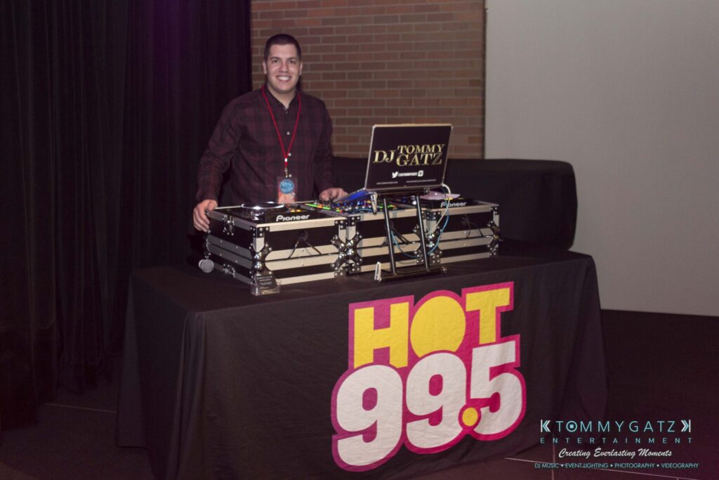 18A0776 - TOMMY GATZ ENTERTAINMENT A DJ stands behind a booth with equipment, featuring a "Hot 99.5" banner.