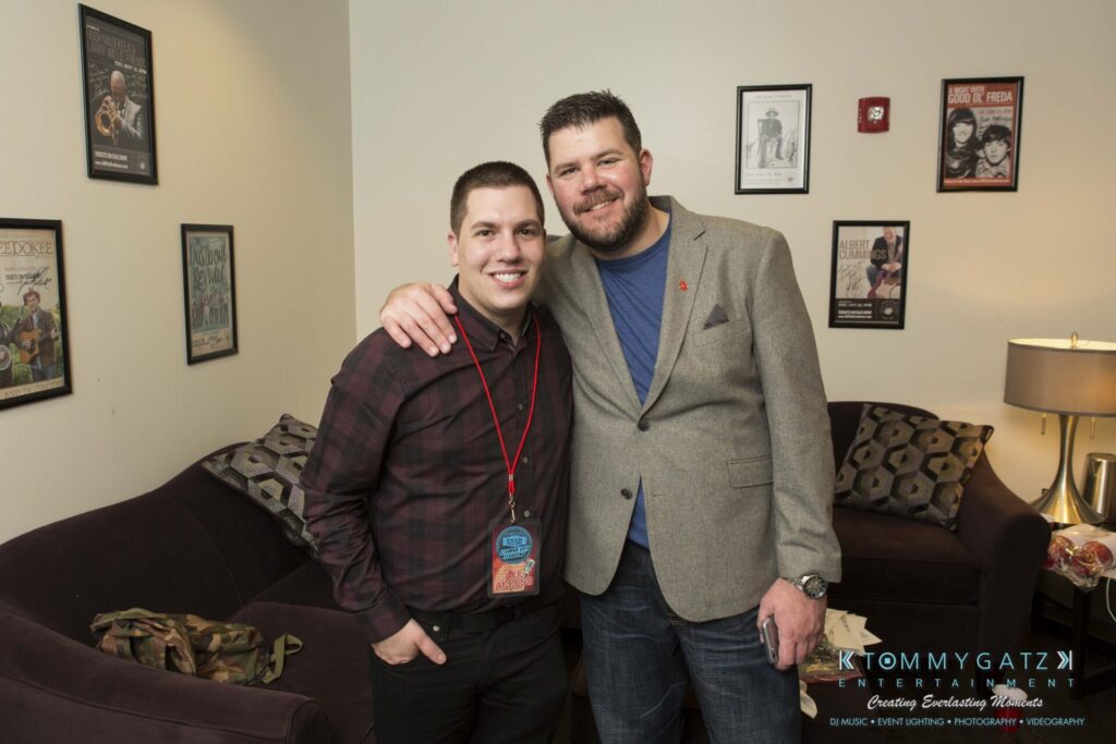18A0785 - TOMMY GATZ ENTERTAINMENT Two men posing together in a living room with framed posters on the wall behind them. One wears a plaid shirt and lanyard, the other a gray blazer. A couch and a standing lamp are visible.