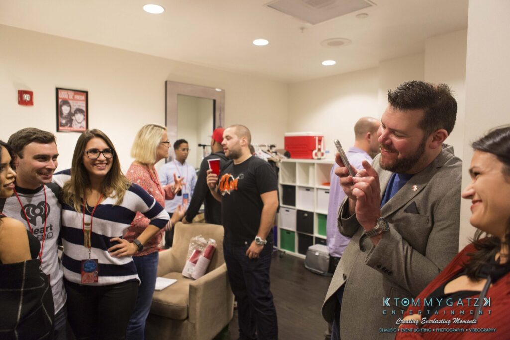 18A0825 - TOMMY GATZ ENTERTAINMENT A group of people socializing indoors, with some holding drinks and one person taking a photo with a smartphone.