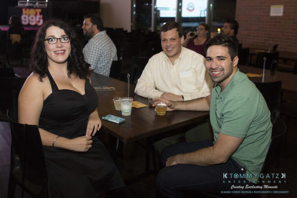 18A0837 - TOMMY GATZ ENTERTAINMENT Three people sitting at a wooden table with drinks, smiling at the camera in a dimly lit venue.