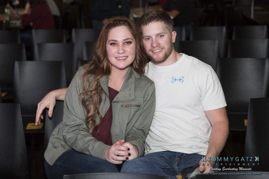 18A0845 - TOMMY GATZ ENTERTAINMENT A person with long hair and a jacket sits next to a person in a white t-shirt, both smiling and seated on black chairs in a casual setting.