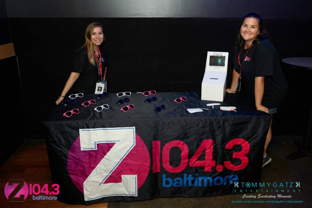 Intern-John-Peer-Pressure-Comedy-Show-Baltimore-7-14-17-Blog-Watermark-14 - TOMMY GATZ ENTERTAINMENT Two people stand behind a table with Z104.3 Baltimore merchandise, including sunglasses and lanyards, at an event.