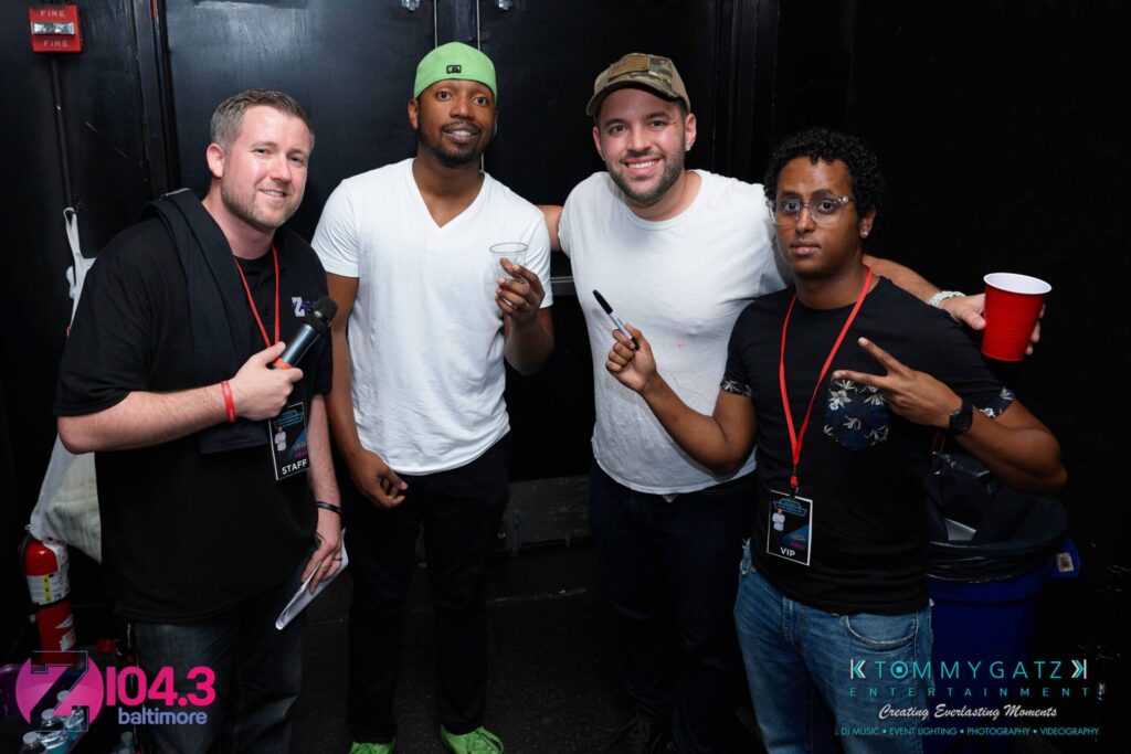 Intern-John-Peer-Pressure-Comedy-Show-Baltimore-7-14-17-Blog-Watermark-16 - TOMMY GATZ ENTERTAINMENT Four men posing together, two holding drinks. They are in a room with black walls, and one wears a green cap. A logo and text about Baltimore's 104.3 radio station is visible in the corner.