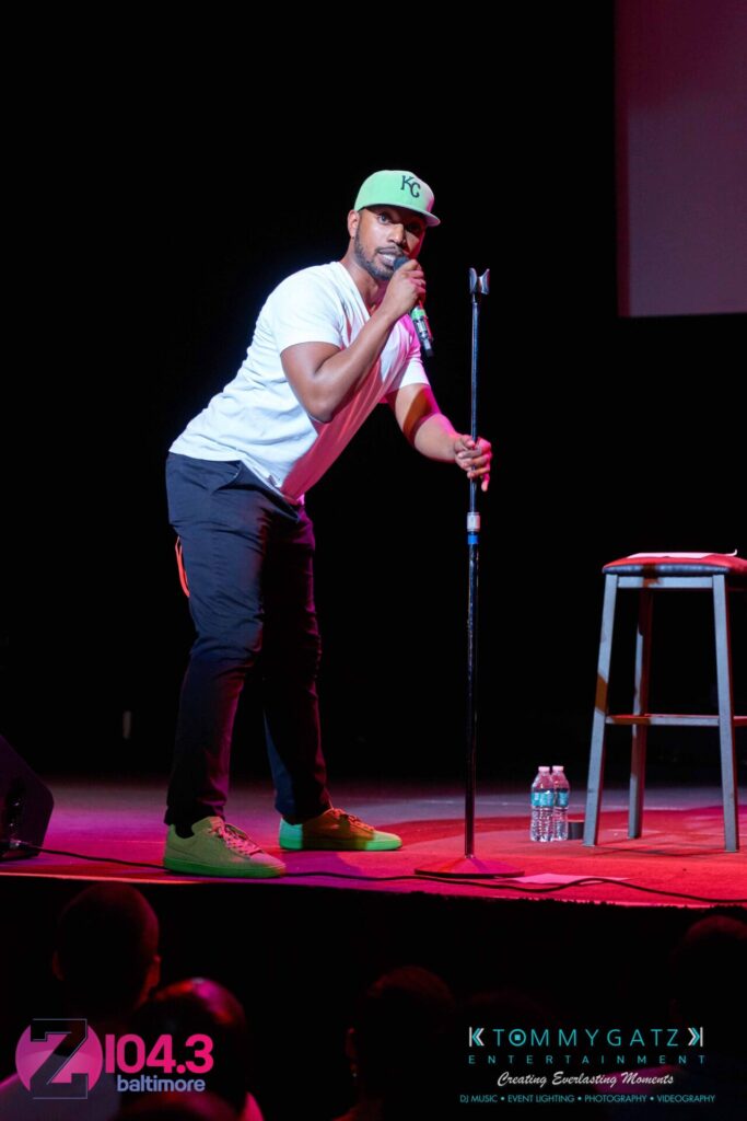 Intern-John-Peer-Pressure-Comedy-Show-Baltimore-7-14-17-Blog-Watermark-18 - TOMMY GATZ ENTERTAINMENT A performer in a white shirt and green cap sings into a microphone on a stage with a stool nearby, under colorful lighting. Logos of "Z104.3 Baltimore" and "Tommy Gatz Entertainment" are visible.