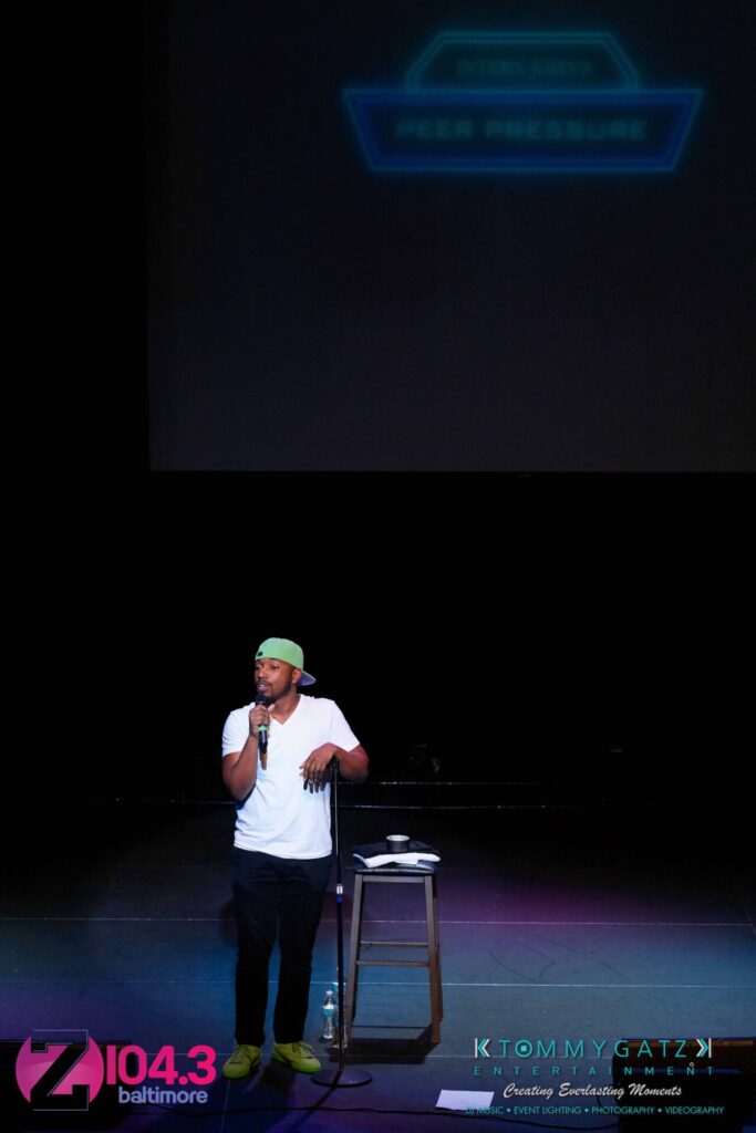 Intern-John-Peer-Pressure-Comedy-Show-Baltimore-7-14-17-Blog-Watermark-19 - TOMMY GATZ ENTERTAINMENT A man wearing a cap is speaking on stage in front of a stool and mic stand, with a Z104.3 Baltimore logo in the bottom corner.