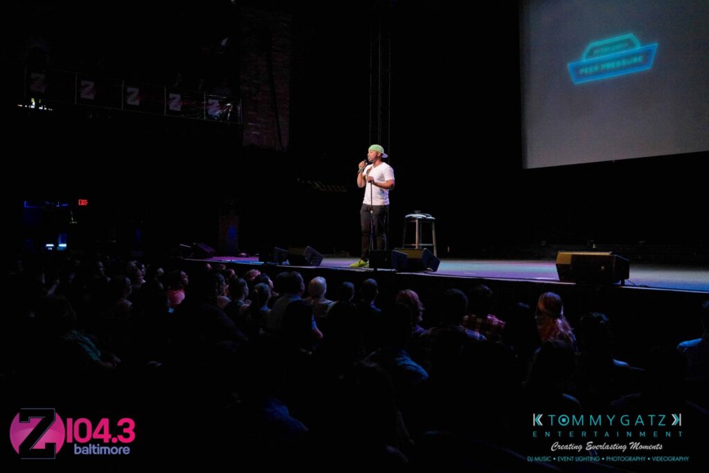 Intern-John-Peer-Pressure-Comedy-Show-Baltimore-7-14-17-Blog-Watermark-21 - TOMMY GATZ ENTERTAINMENT A person stands on stage performing in front of a seated audience, with logos and company names visible on stage and the foreground.