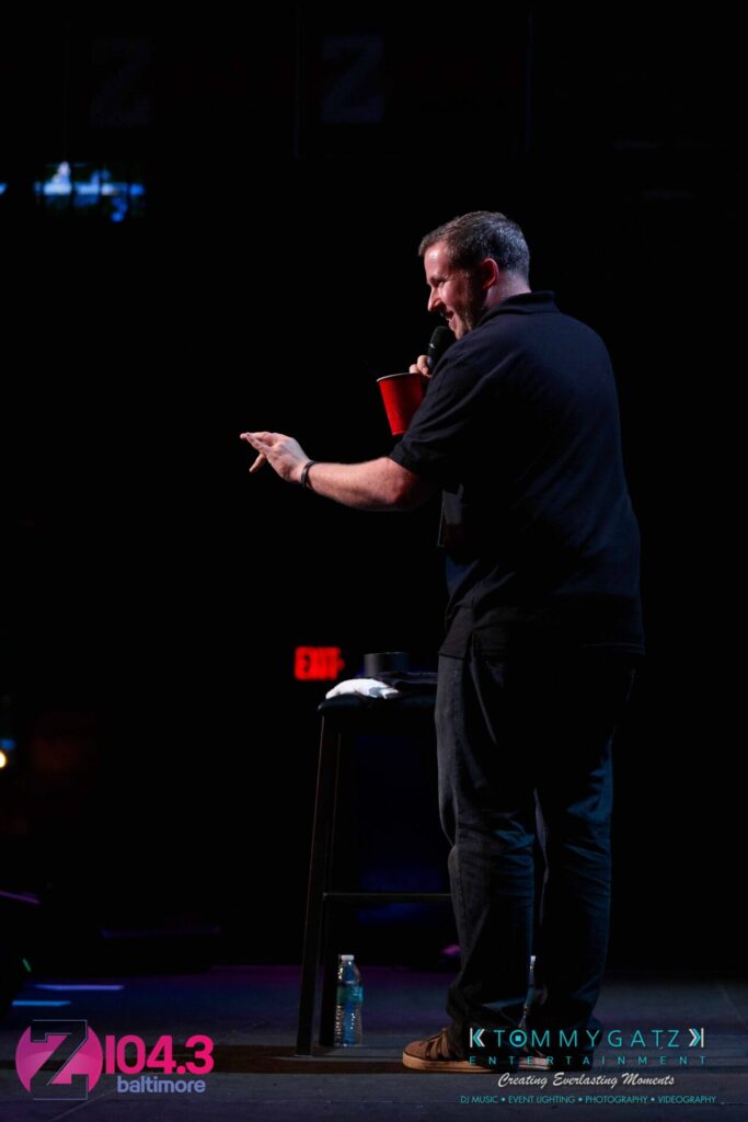 Intern-John-Peer-Pressure-Comedy-Show-Baltimore-7-14-17-Blog-Watermark-22 - TOMMY GATZ ENTERTAINMENT A man stands on stage holding a red cup and microphone, facing an audience. There's a stool and bottle of water nearby.