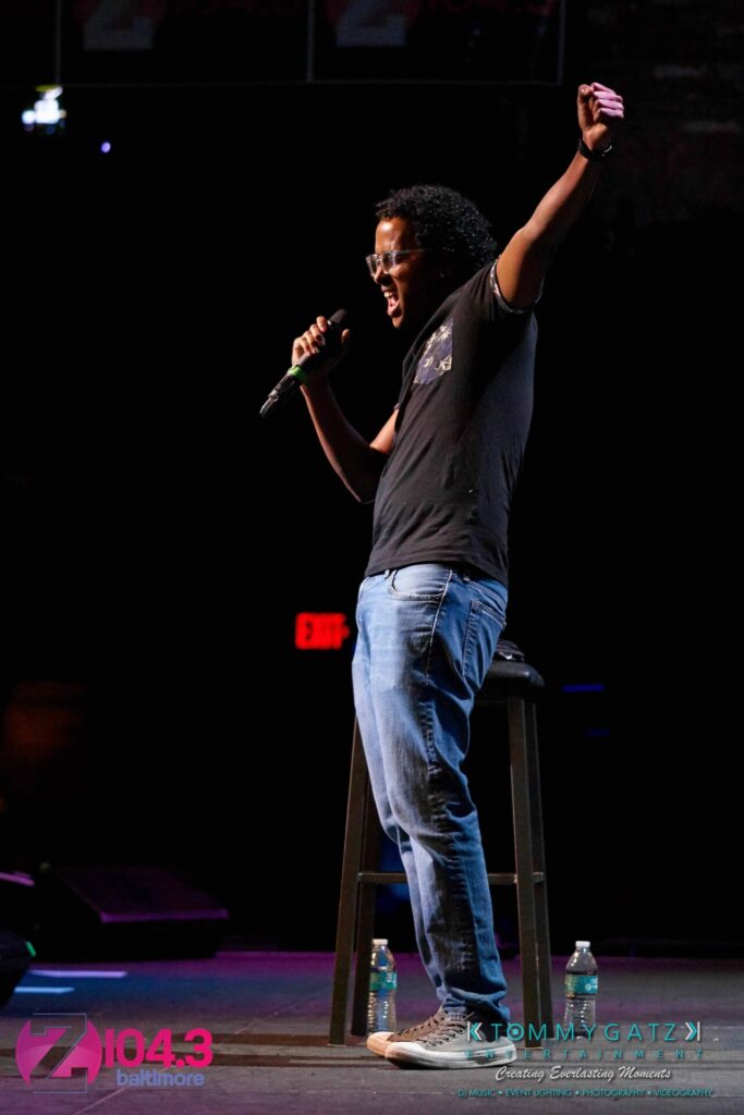 Intern-John-Peer-Pressure-Comedy-Show-Baltimore-7-14-17-Blog-Watermark-24 - TOMMY GATZ ENTERTAINMENT A man stands on stage holding a microphone, gesturing passionately. He is next to a stool with two water bottles on the floor. Banners for Z104.3 Baltimore are in the background.