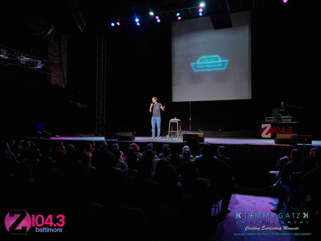Intern-John-Peer-Pressure-Comedy-Show-Baltimore-7-14-17-Blog-Watermark-25 - TOMMY GATZ ENTERTAINMENT Comedian performing on stage with a microphone, audience seated in front. Bright stage lights and a screen displaying text behind the performer. Radio station logo visible on stage.