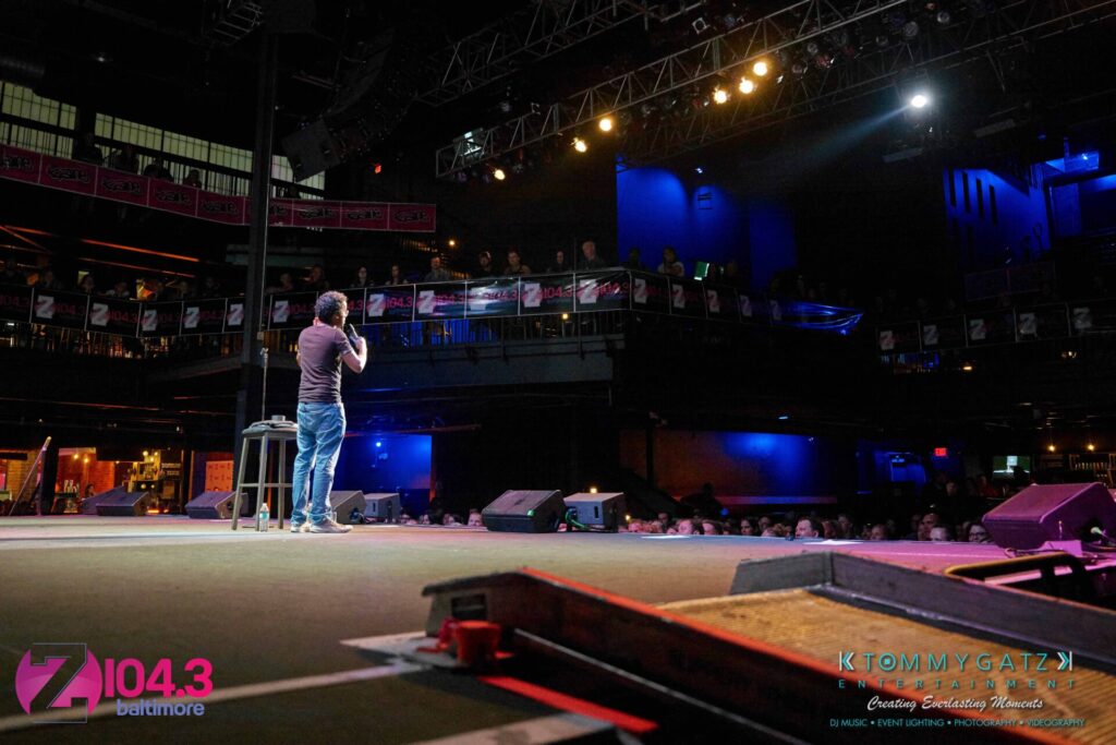 Intern-John-Peer-Pressure-Comedy-Show-Baltimore-7-14-17-Blog-Watermark-26 - TOMMY GATZ ENTERTAINMENT Comedian performing on stage at a venue with a seated audience. Stage lights are focused on the performer, and a large banner in the background displays the radio station Z104.3 Baltimore.