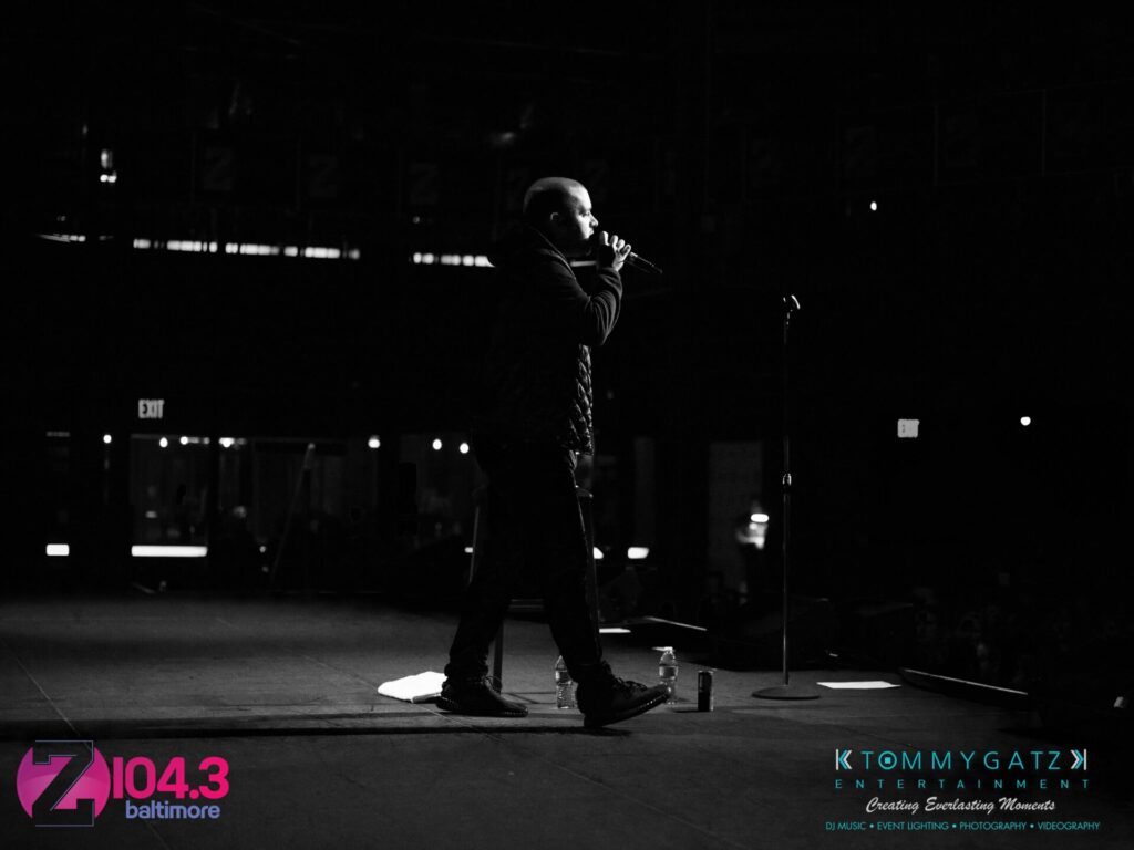 Intern-John-Peer-Pressure-Comedy-Show-Baltimore-7-14-17-Blog-Watermark-28 - TOMMY GATZ ENTERTAINMENT A person on stage holds a microphone, performing in front of a dark audience. Logos for Z104.3 Baltimore and Tommy Gatz Entertainment are visible.