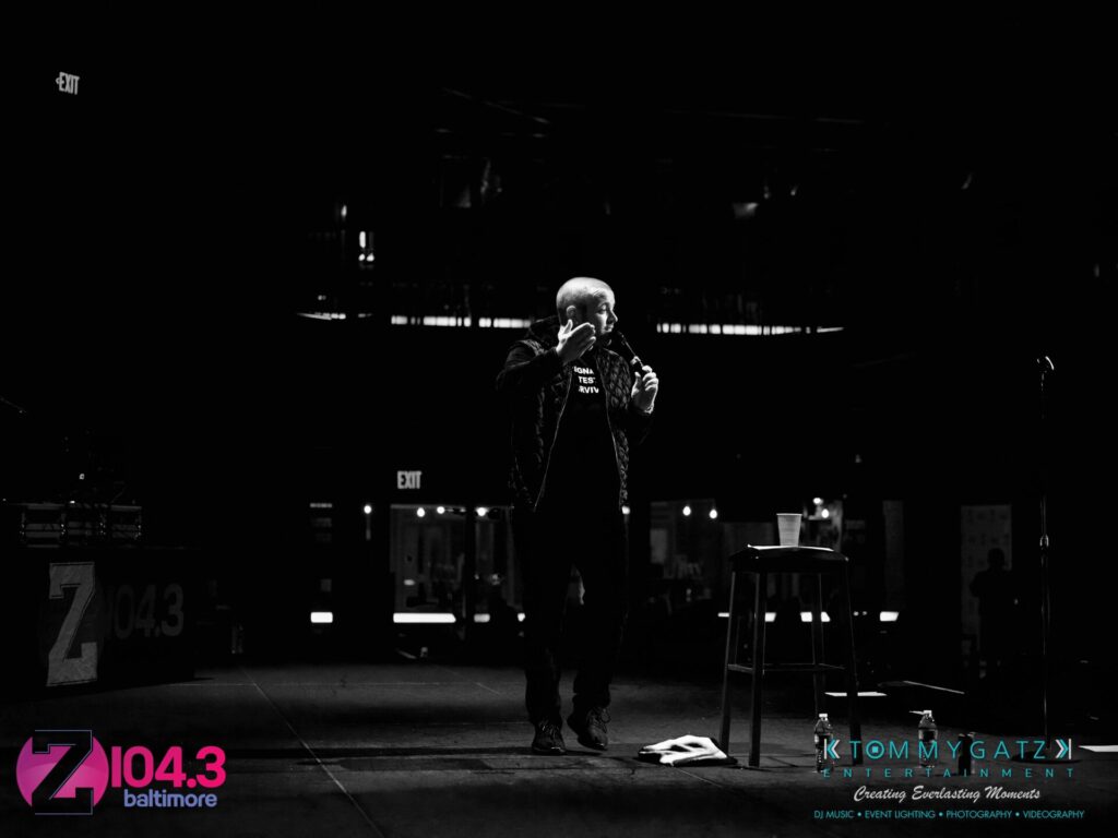 Intern-John-Peer-Pressure-Comedy-Show-Baltimore-7-14-17-Blog-Watermark-29 - TOMMY GATZ ENTERTAINMENT A person on stage is talking into a microphone. The stage is set up with a stool and a water bottle. Z104.3 and Tommy Gatz Entertainment logos are visible on the image.