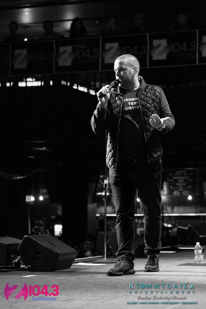 Intern-John-Peer-Pressure-Comedy-Show-Baltimore-7-14-17-Blog-Watermark-32 - TOMMY GATZ ENTERTAINMENT A person stands on stage holding a microphone, speaking to an audience. The background features branding for a radio station.