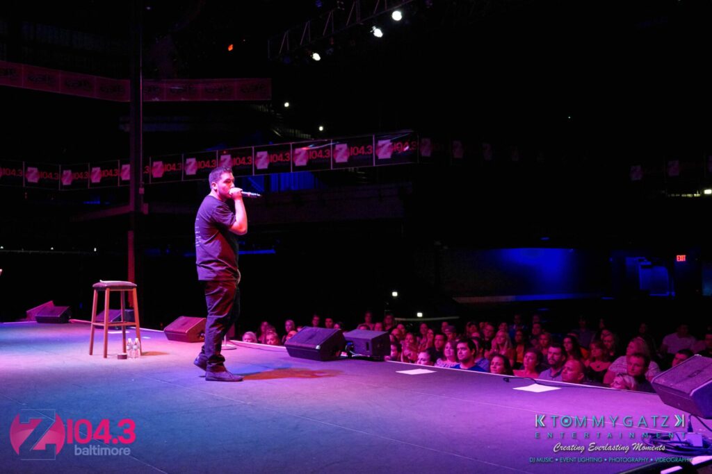Intern-John-Peer-Pressure-Comedy-Show-Baltimore-7-14-17-Blog-Watermark-36 - TOMMY GATZ ENTERTAINMENT Comedian performing on stage in front of a seated audience, with a stool beside him. Stage banners and lighting visible.