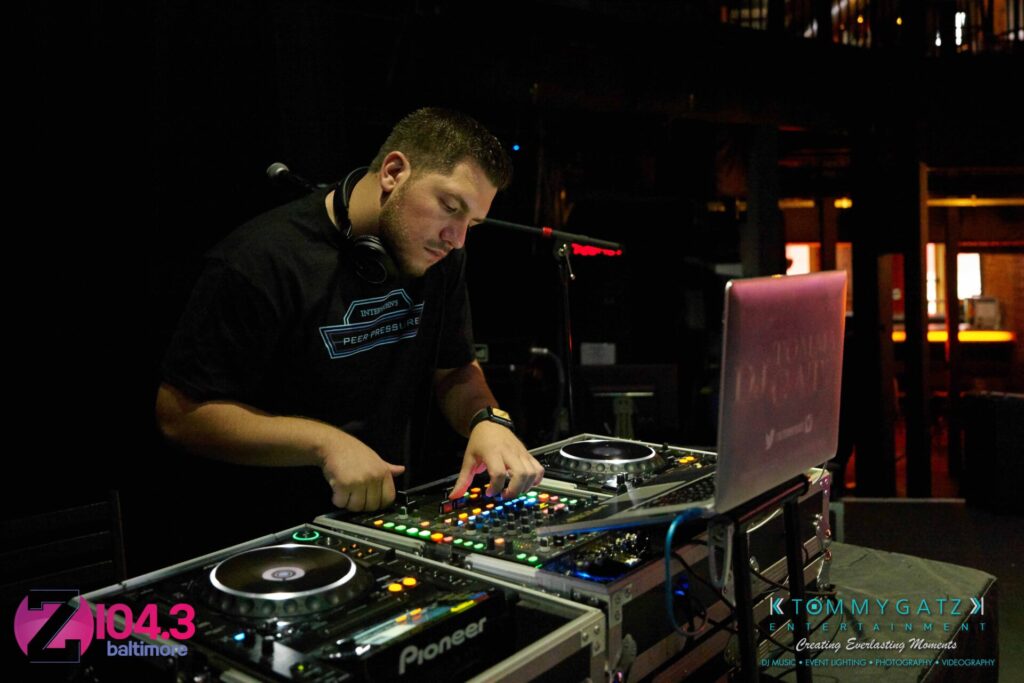 Intern-John-Peer-Pressure-Comedy-Show-Baltimore-7-14-17-Blog-Watermark-4 - TOMMY GATZ ENTERTAINMENT A DJ with headphones operates music equipment on a dimly lit stage. Multiple logos are visible in the scene.