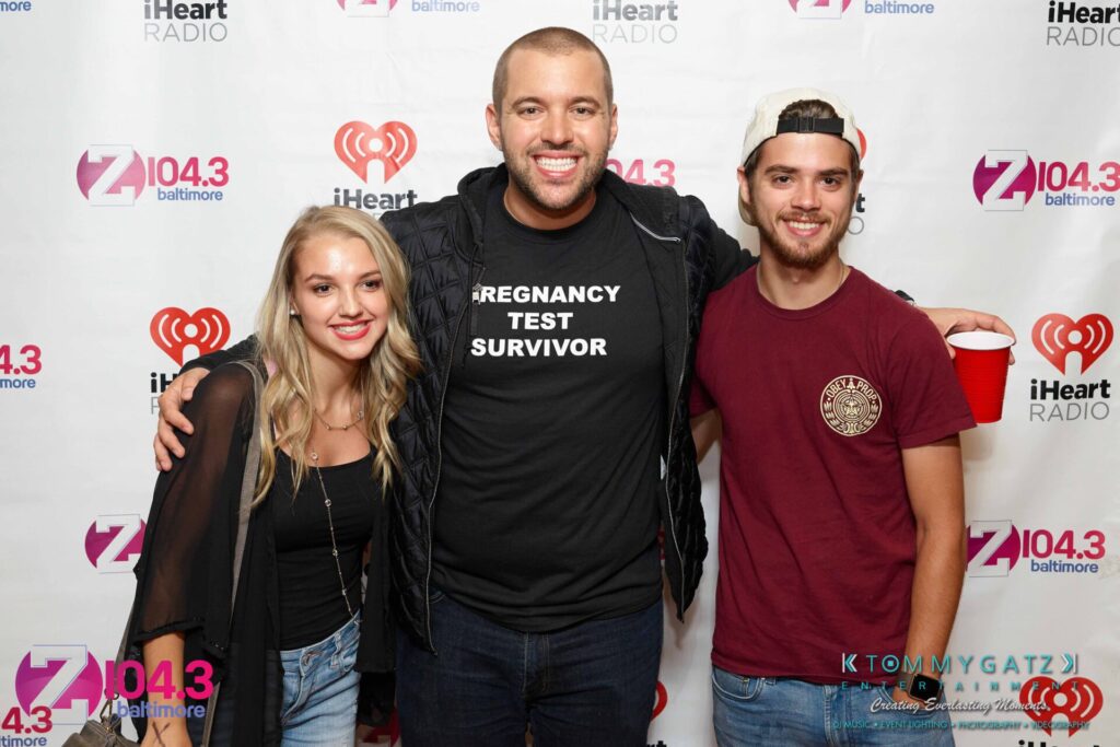 Intern-John-Peer-Pressure-Comedy-Show-Baltimore-7-14-17-Blog-Watermark-42 - TOMMY GATZ ENTERTAINMENT Three people pose in front of a backdrop with logos, including iHeart Radio and Z104.3. The center person wears a shirt reading "Pregnancy Test Survivor.