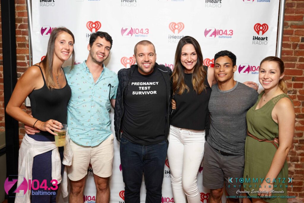 Intern-John-Peer-Pressure-Comedy-Show-Baltimore-7-14-17-Blog-Watermark-43 - TOMMY GATZ ENTERTAINMENT A group of six people posing together in front of a backdrop. One person in the center wears a "Pregnancy Test Survivor" shirt. Logos for Z104.3 and iHeart Radio are visible.