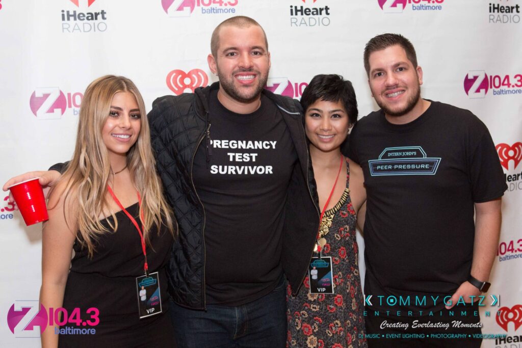 Intern-John-Peer-Pressure-Comedy-Show-Baltimore-7-14-17-Blog-Watermark-48 - TOMMY GATZ ENTERTAINMENT Four people posing in front of a step-and-repeat banner with logos from iHeart Radio and Z104.3 Baltimore. One person holds a red cup. Two are wearing VIP lanyards.
