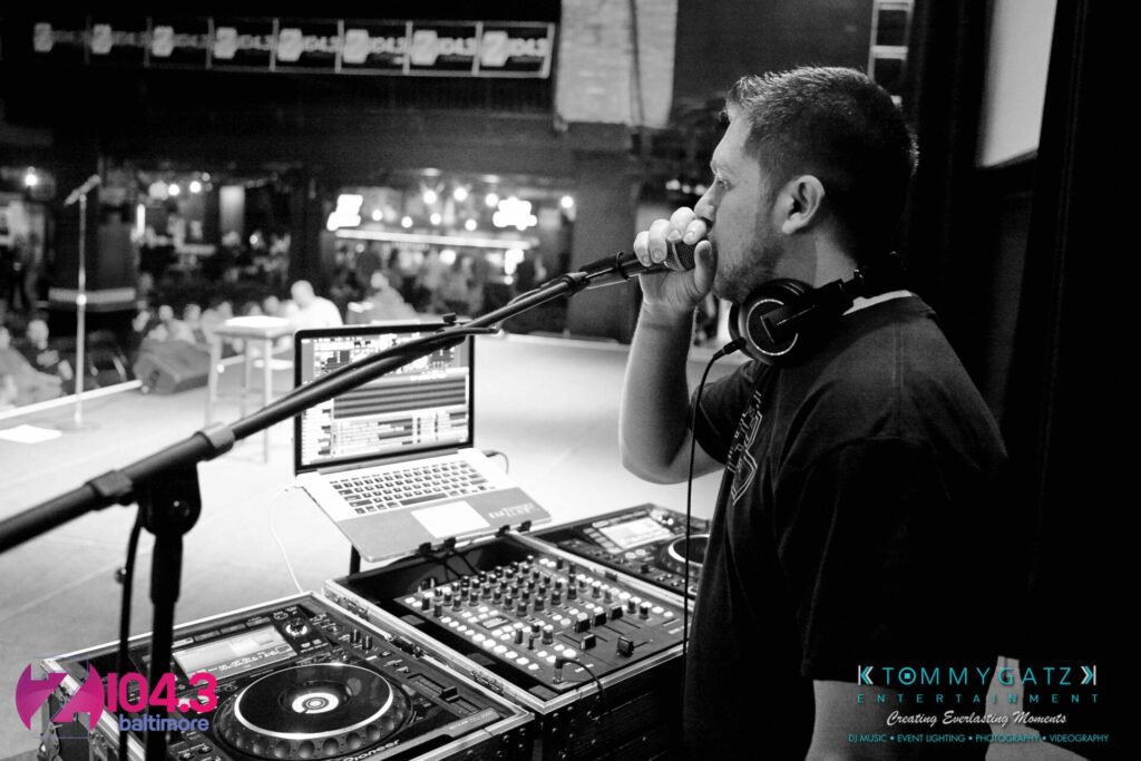 Intern-John-Peer-Pressure-Comedy-Show-Baltimore-7-14-17-Blog-Watermark-6 - TOMMY GATZ ENTERTAINMENT A DJ is performing on stage, holding a microphone, with music equipment in front of him and a laptop open to his right.