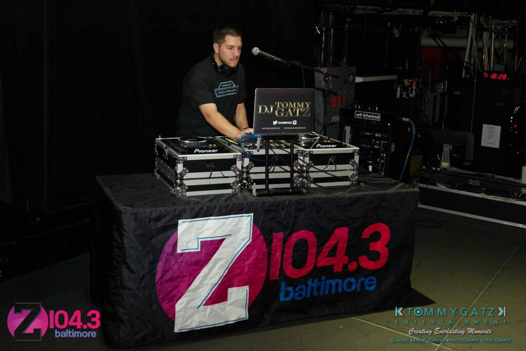 Intern-John-Peer-Pressure-Comedy-Show-Baltimore-7-14-17-Blog-Watermark-7 - TOMMY GATZ ENTERTAINMENT A DJ stands behind a table with audio equipment, branded with "Z104.3 Baltimore" and "DJ Tommy Gatz." The setting appears to be a dimly lit indoor venue.