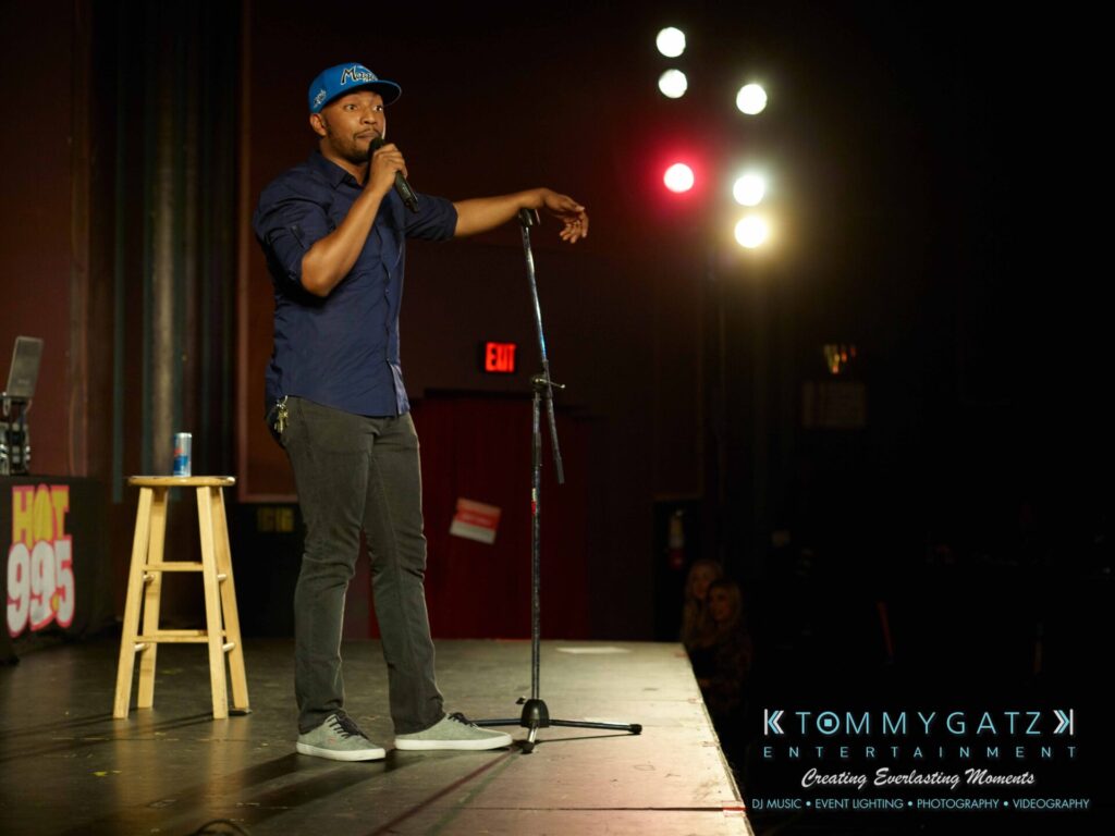 Intern-John-Peer-Pressure-Comedy-Tour-Arlington-7-21-17-low-res-Watermark-51 - TOMMY GATZ ENTERTAINMENT Comedian performing on stage with a microphone, stool, and drink can beside him. Bright lights illuminate the scene, and an audience is partially visible. Logo for Tommy Gatz Entertainment appears.
