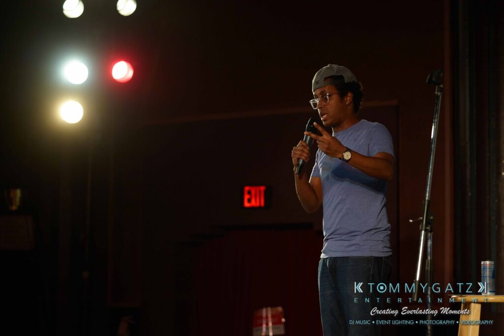 Intern-John-Peer-Pressure-Comedy-Tour-Arlington-7-21-17-low-res-Watermark-60 - TOMMY GATZ ENTERTAINMENT A person in a blue shirt and cap speaks into a microphone on stage, under bright lights, with "Tommy Gatz Entertainment" text overlay.