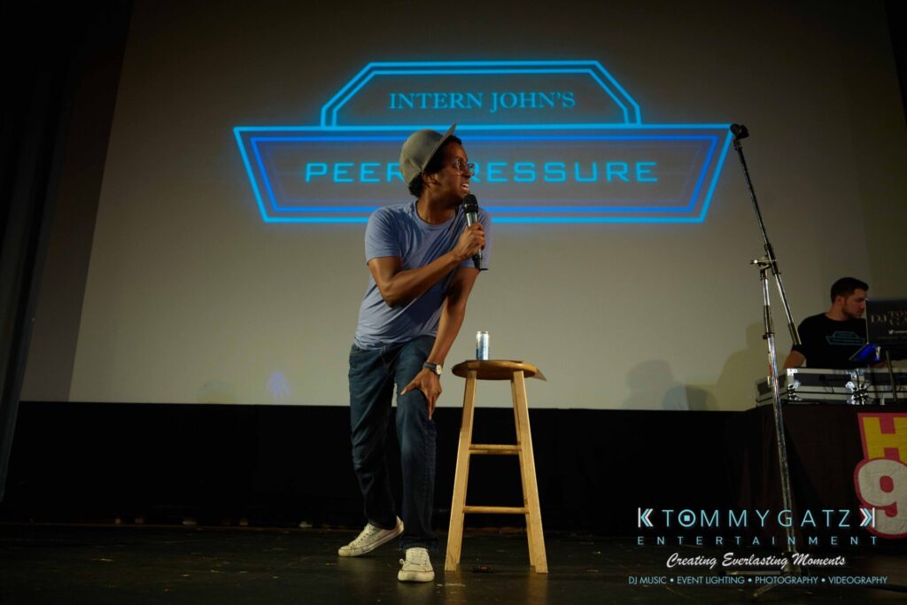 Intern-John-Peer-Pressure-Comedy-Tour-Arlington-7-21-17-low-res-Watermark-64 - TOMMY GATZ ENTERTAINMENT Person speaking into a microphone on stage next to a wooden stool, with a projected sign reading "Intern John's Peer Pressure" in the background.