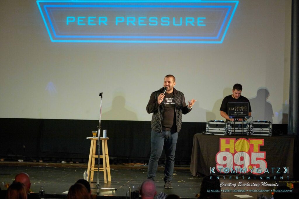 Intern-John-Peer-Pressure-Comedy-Tour-Arlington-7-21-17-low-res-Watermark-77 - TOMMY GATZ ENTERTAINMENT A man speaks into a microphone on stage with a DJ and a sign reading "Peer Pressure" above them.