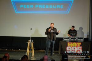 A man speaks into a microphone on stage with a DJ and a sign reading "Peer Pressure" above them.