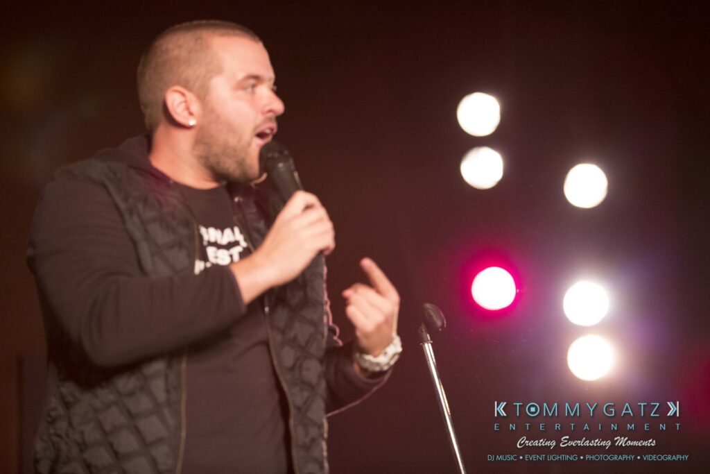 Intern-John-Pressure-Comedy-Tour-ARLINGTON-BLOG-WM-130-of-254 - TOMMY GATZ ENTERTAINMENT A man on stage holds a microphone, gesturing while speaking. Bright lights shine in the background.
