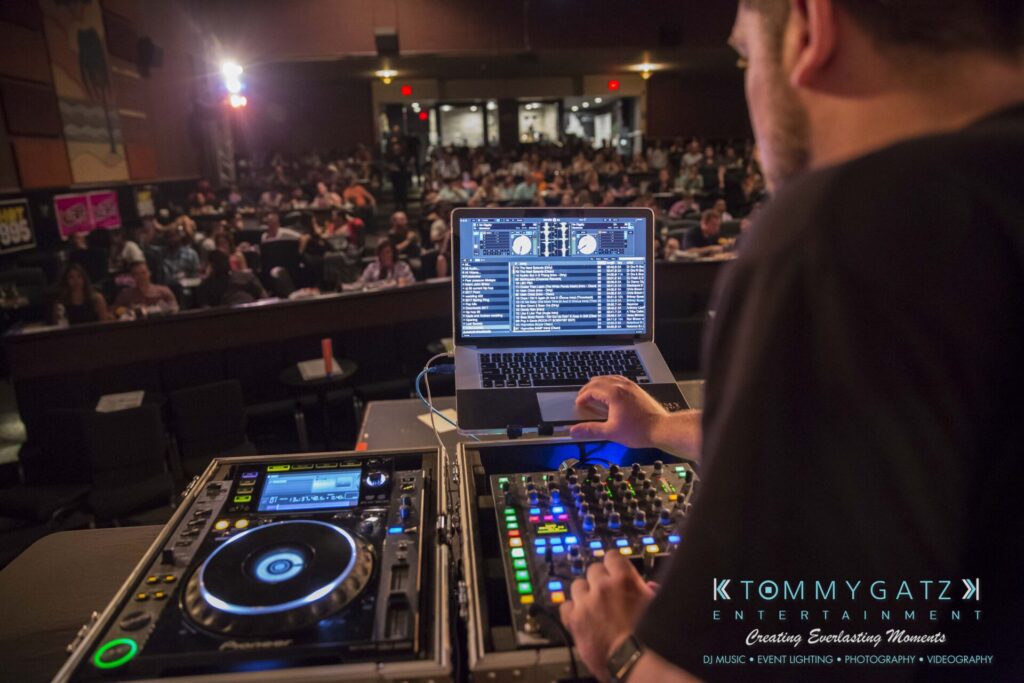 Intern-John-Pressure-Comedy-Tour-ARLINGTON-BLOG-WM-17-of-254 - TOMMY GATZ ENTERTAINMENT A DJ stands at a console with turntables and a laptop, mixing music for an engaged audience in a dimly lit venue.