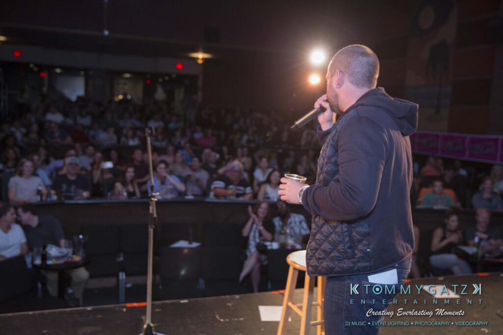 Intern-John-Pressure-Comedy-Tour-ARLINGTON-BLOG-WM-214-of-323 - TOMMY GATZ ENTERTAINMENT A person with a microphone addresses a seated audience in a dimly lit venue. A spotlight shines from the stage, and there are visible logos at the bottom of the image.