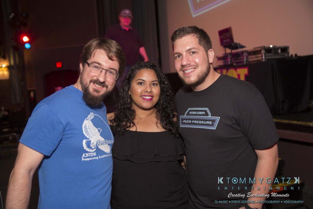 Intern-John-Pressure-Comedy-Tour-ARLINGTON-BLOG-WM-238-of-323 - TOMMY GATZ ENTERTAINMENT Three people smiling in a dimly lit indoor event space, standing in front of a screen and DJ equipment.