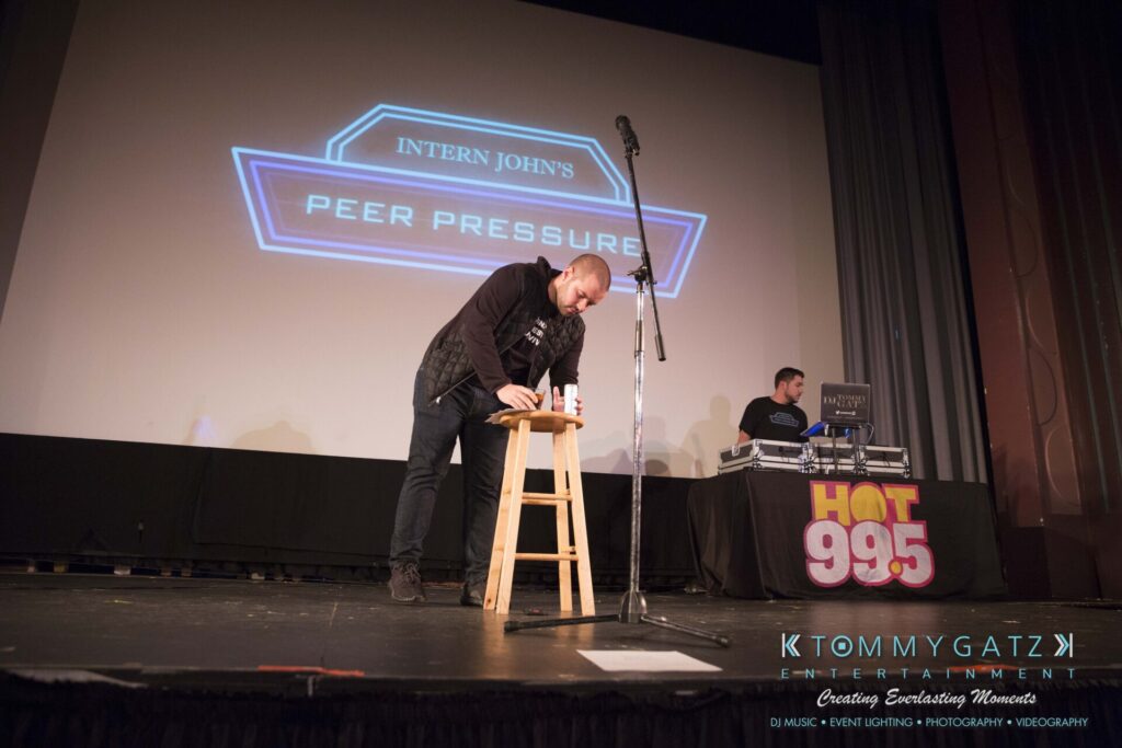 Intern-John-Pressure-Comedy-Tour-ARLINGTON-BLOG-WM-94-of-254 - TOMMY GATZ ENTERTAINMENT A man is standing and writing on a notepad on a stool on stage, with a microphone next to him and a background screen displaying "Intern John's Peer Pressure.