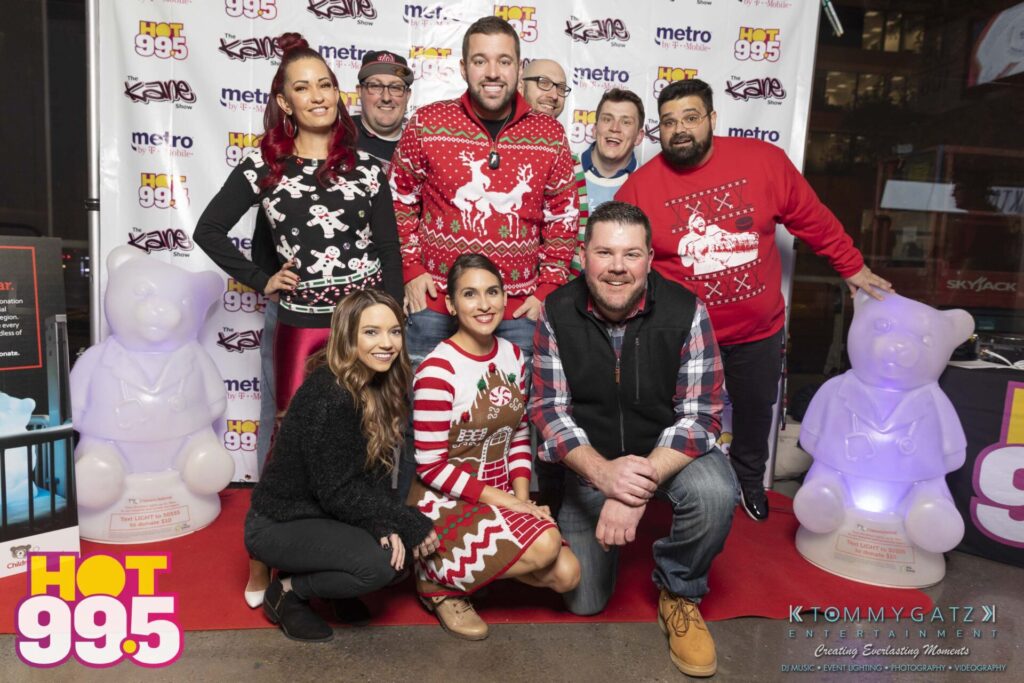 Jingle-Ball-2018-101 - TOMMY GATZ ENTERTAINMENT A group of people in festive attire pose together at a Hot 99.5 event, standing on a red carpet with branded banners and ice sculptures in the background.