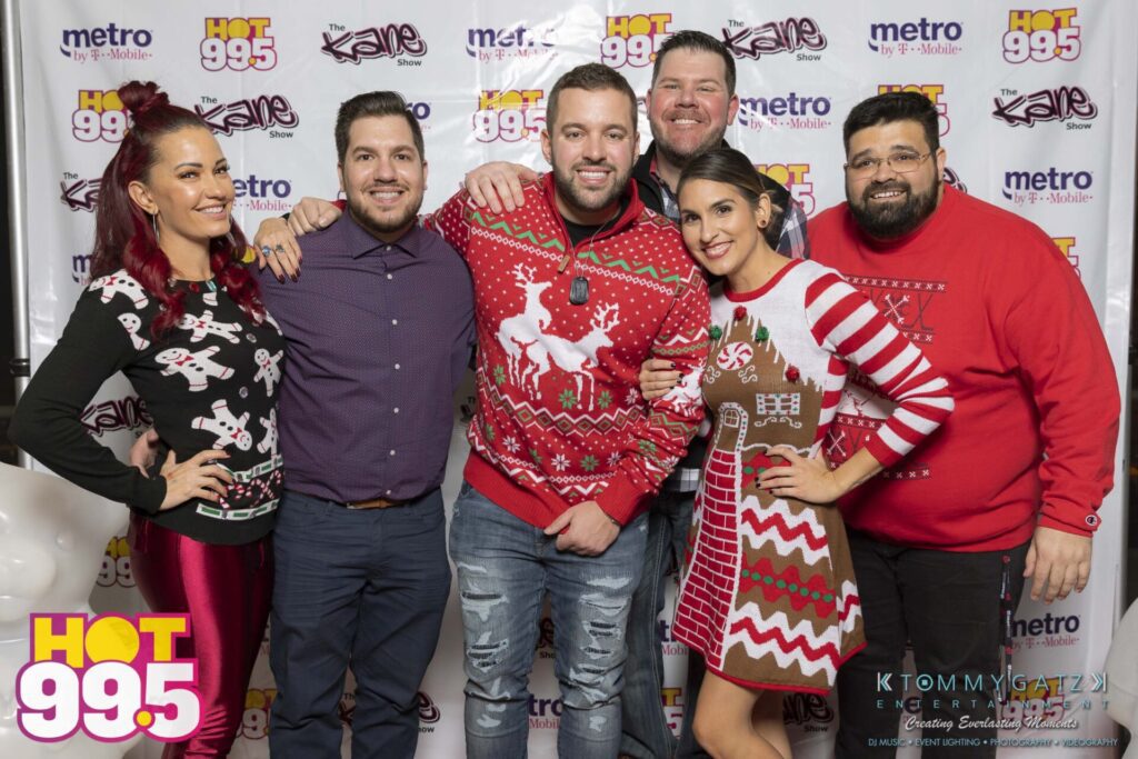 A group of people posing in front of a Hot 99.5 backdrop, wearing festive holiday outfits.